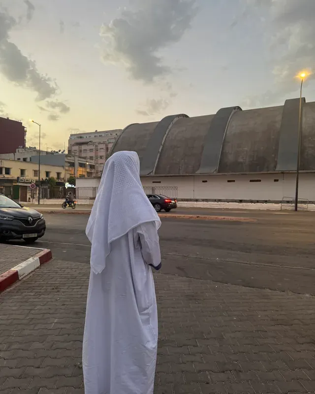Une rue de Casablanca au crépuscule. Au premier plan, une silhouette de dos en djellaba blanche traverse le trottoir pavé ; en face, la longue halle du marché aux toits arrondis, quelques voitures et un lampadaire allumé sous un ciel doré.